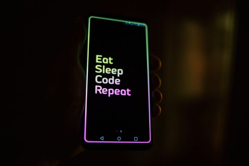 A hand holding a smartphone with a screen displaying the text 'Eat Sleep Code Repeat' in neon colors.
